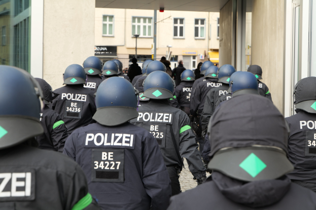 Eine Gruppe von Polizisten in Uniform und Helmen steht vor einem Gebäude, im Hintergrund sind Menschen, Pfähle und andere Gebäude zu sehen.