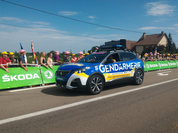 Ein Polizeiwagen mit einem Banner fährt an einer Menge vorbei, die Fahnen schwenkt und Mötzen trägt, mit Bäumen, Gebäuden und Wolken im Hintergrund; Text unten lautet "Gendarmerie de la Tour de France."