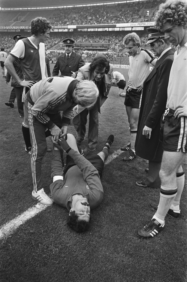 Schwarzes und weißes Bild eines Fußballspielers auf dem Boden umgeben von einer Gruppe von Männern, mit einer Zuschauermenge im Hintergrund eines Stadions.