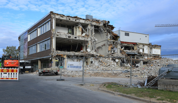 Ein großes Gebäude wird in der Mitte einer Straße abgerissen, umgeben von einem Metallzaun, mit Fahrzeugen, Gras, einem Schild, einer Fahne, Pfählen, einem Kran, Bäumen und einem bewölkten Himmel.