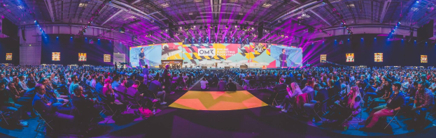 Eine große Zuschauermenge sitzt in Stühlen gegenüber einer hell erleuchteten Bühne mit Bildschirmen, auf der mehrere Menschen stehen während eines Online-Marketing-Events.