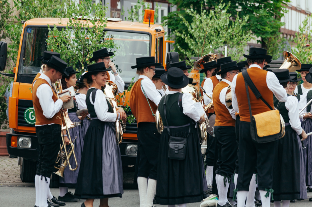 Eine Gruppe von Menschen in traditioneller bayrischer Tracht marschiert die Straße entlang, spielt Instrumente und trägt Taschen, mit Bäumen, Gebäuden und einem geparkten Fahrzeug im Hintergrund.