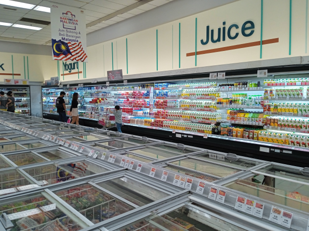 Ein hell erleuchteter Saftladen in einem Supermarkt, der eine Auswahl an Flaschengetränken, frisches Gemüse und andere Artikel in Regalen anbietet, mit ein paar Menschen, die durch die Gänge schlendern, und Deckenlampen, die den Bereich beleuchten.