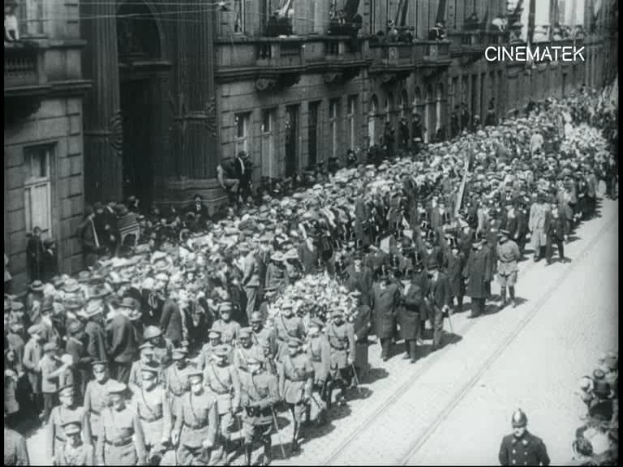 Schwarzes und weißes Foto einer Parade mit einer großen Menge von Menschen, die Gewehre tragen und an einem Gebäude vorbeimarschieren, mit einem Wasserzeichen in der oberen rechten Ecke.