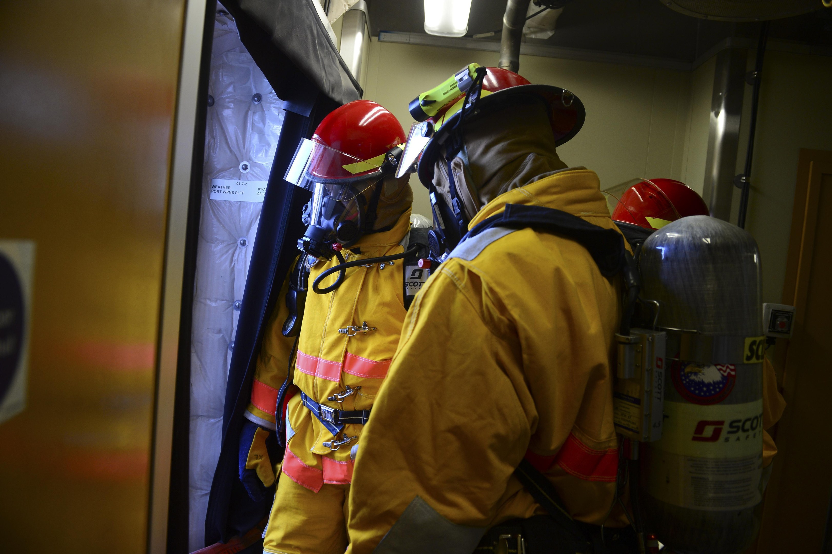 Feuerwehrleute in gelben Uniformen und roten Helmen stehen in einer Gruppe rechts neben Zylindern und links neben einer Tür, mit einer Wand und Licht im Hintergrund.