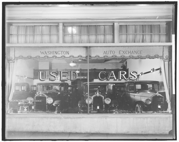 Schwarze und weiße Fotografie eines Gebrauchtwagenhändlers in Washington, D.C., mit mehreren Autos durch die Glasscheibe sichtbar, Text an der Wand und beleuchteter Beleuchtung.