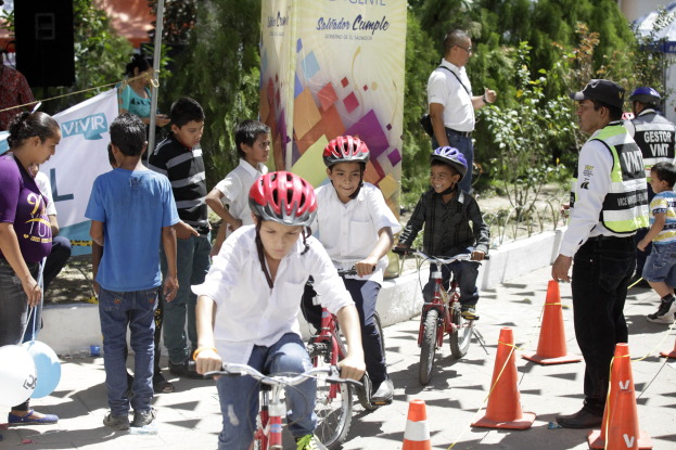Eine Gruppe von Kindern, die auf Fahrraedern eine Strasse entlangfahren, mit Verkehrskegeln, einige tragen Helme, andere stehen in der Nahe, mit einer Fahne, Baeumen und Gebaeuden im Hintergrund.