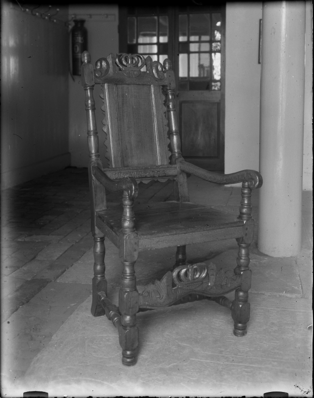 Schwarzes und weißes Foto eines alten Holzstuhls in einem Raum mit einer Tür und einer Säule im Hintergrund.