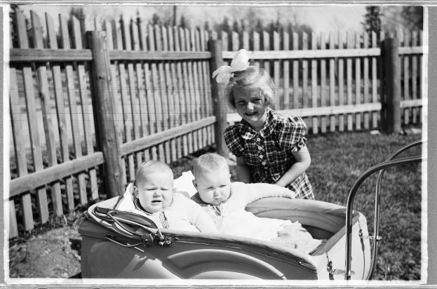 Schwarzes und weißes Foto von drei Kindern in einem Auto, mit einem Mädchen, das im Vordergrund steht und lächelt, einem Zaun und Bäumen im Hintergrund und grasbewachsenem Boden.