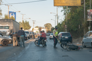 Eine Gruppe von Menschen steht um ein verunglücktes Motorrad am Rande einer Straße mit mehreren Fahrzeugen, darunter ein Lastwagen, und einem Hintergrund aus Bäumen, Masten, Lichtern und Schildern unter dem Himmel.