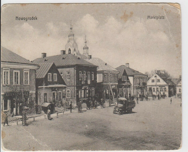 Schwarz-weiß-Fotografie einer Stadtstraße in Nowogrodeek, Deutschland, mit Gebäuden, Menschen, Wagen, Pfählen, Bäumen und Himmel, mit der Beschriftung "Marktplatz in Nowogrodeek, Deutschland" oben und unten.