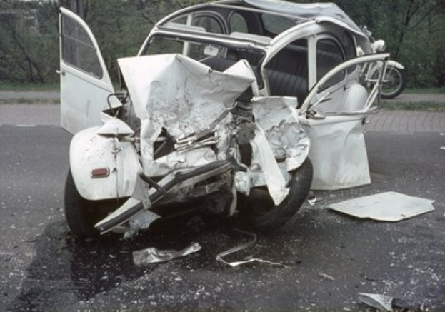 Schwer beschädigte Frontpartie eines Wagens, der auf der Straße neben der Fahrbahn steht, mit Bäumen und einem Gebäude im Hintergrund und einem weiteren Fahrzeug in der Ferne.