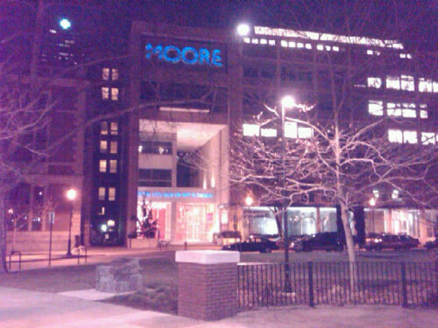 Außenansicht eines gebäudeähnlichen Krankenhauses mit parkenden Fahrzeugen davor, umgeben von Bäumen und einem Zaun.