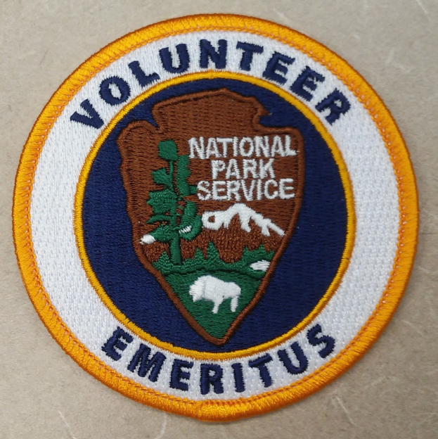 Runder Patch mit blauem Rand und weißem Hintergrund, der den fetten schwarzen Text "Volunteer National Park Service Emeritus" in der Mitte eines gelben Kreises zeigt.