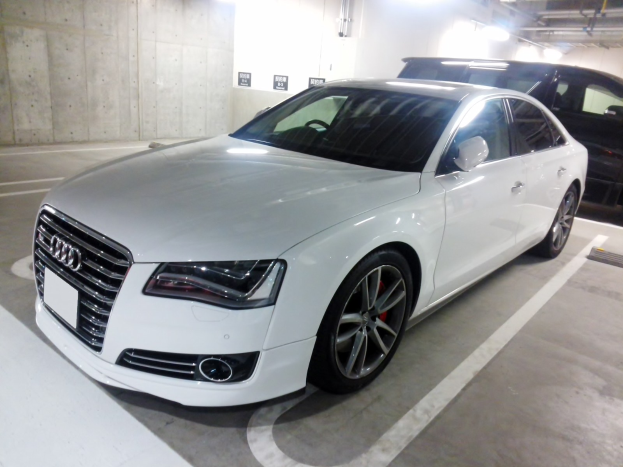 Weißer Audi S8 auf einem Parkplatz neben einem schwarzen Auto geparkt, mit Wänden, Deckenrohren und Leuchten im Hintergrund.