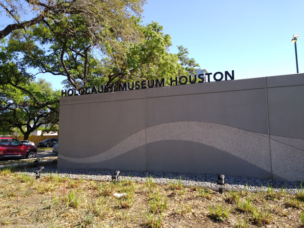 Das Holocaust-Museum in Houston, Texas, mit einer textbedeckten Wand, Steinen, Gras, Pflanzen, Bäumen, Fahrzeugen auf der Straße und einem Laternenpfahl.