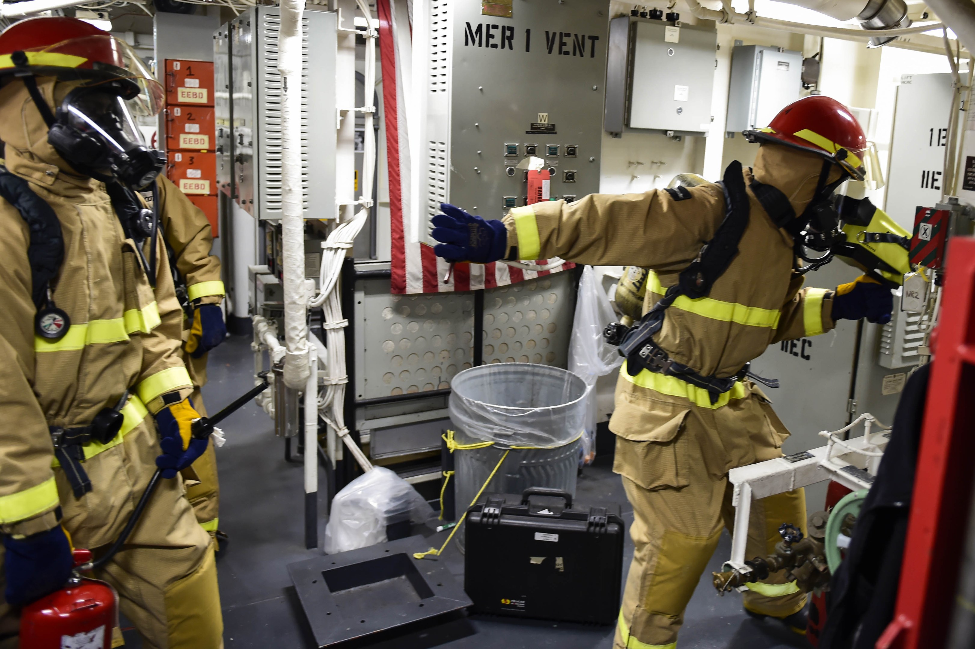 Drei Feuerwehrleute in Schutzausrüstung stehen in einem Raum mit einem Feuerlöscher links, einer Maschine rechts und verschiedenen Gegenständen und Rohren im Hintergrund.