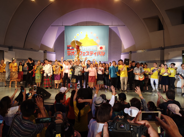 Gruppe von Menschen auf einer Bühne während einer Festival-Eröffnung, einige halten Kameras oder Handys und andere Musikinstrumente, mit einem Banner und Deckenbeleuchtung im Hintergrund.