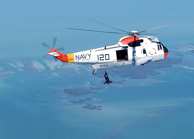 Hubschrauber fliegt über Wasser mit einer Person darunter und Himmel im Hintergrund, als Rettungshubschrauber gekennzeichnet.