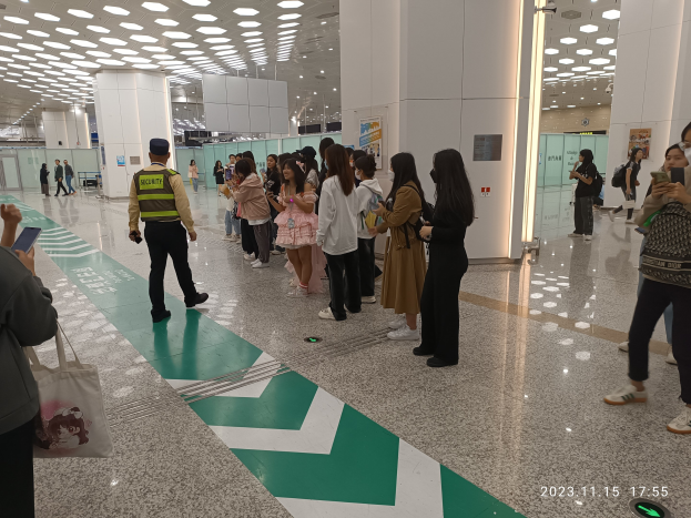 Eine Gruppe von Menschen, die in einer Reihe an einem Flughafen-Sicherheitscheck-in stehen, einige halten Taschen und Mobiltelefone, mit Säulen und Deckenlampen im Hintergrund.