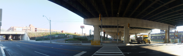 Ein Lastwagen fährt unter einer Brücke auf einer städtischen Straße, die von Gebäuden, Polen, Straßenlaternen, Verkehrszeichen, Fahrzeugen und Grünflächen gesäumt ist, unter einem klaren blauen Himmel.