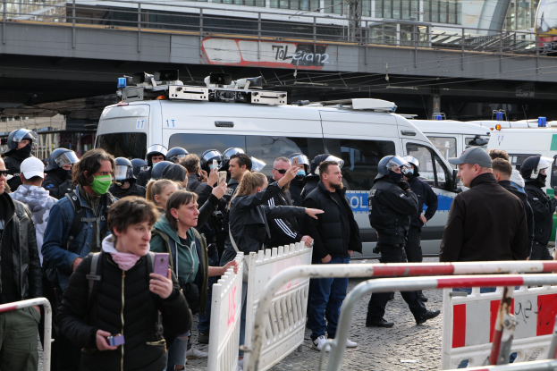 Eine Gruppe von Menschen steht hinter Barrieren und schaut auf eine Reihe von Polizeiwagen, einige tragen Helme und halten Telefone, mit einer Brücke und Gebäuden im Hintergrund während einer Demonstration in Berlin.