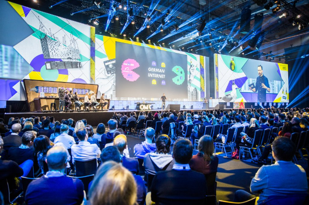 Eine große Menschenmenge sitzt auf Stühlen, die einer Bühne zugewandt sind, mit einem Tisch voller Gegenstände links und Bildschirmen und Lichtern im Hintergrund.