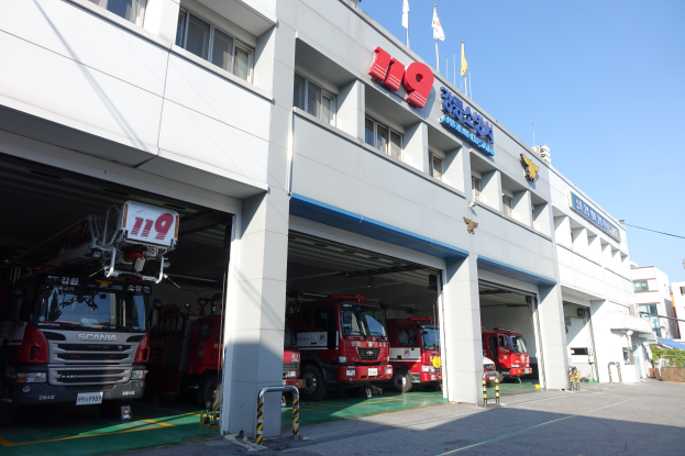 Feuerwache mit mehreren Feuerwehrautos vor der Tür, mit Fenstern, Säulen, Namensschildern und Fahnenmasten im Vordergrund, vor anderen Gebäuden unter einem klaren blauen Himmel.