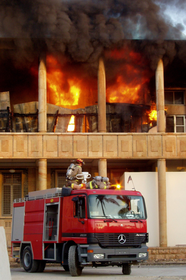 Ein Feuerwehrauto steht auf einer Stra├če vor einem in Flammen stehenden und rauchenden Geb├Ąude, mit einer Wand links daneben.