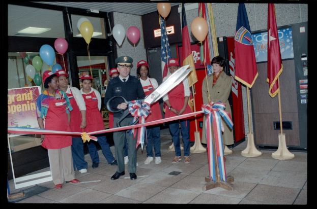 Eine Gruppe von Menschen vor einem Laden mit einem Mann, der eine Schere hält und ein Band durchschneidet, umgeben von Flaggen, Ballons, Texttafeln, Glastüren und Deckenlampen.