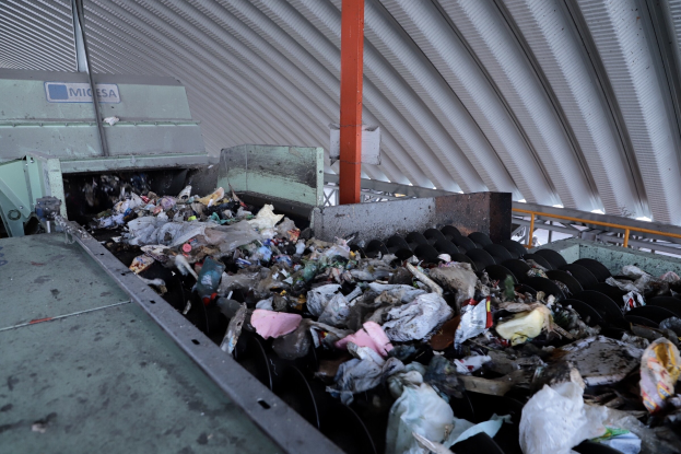 Großer Haufen Müll auf einem Förderband in einer Recyclinganlage mit einer Maschine im Vordergrund und einem Metalldach darüber.