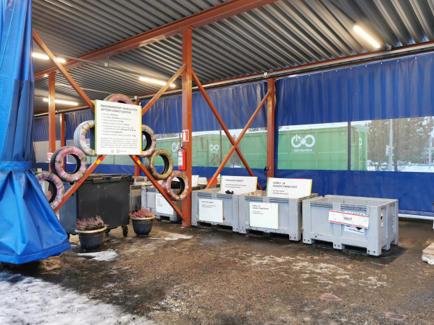Großes Lagerinnere mit verschiedenen Reifen, Containern, Topfpflanzen, bedruckten Platten, Pfählen, Beleuchtung, einem blauen Tuch und anderen Gegenständen, mit Bäumen, Fahrzeugen und Schnee im Hintergrund.