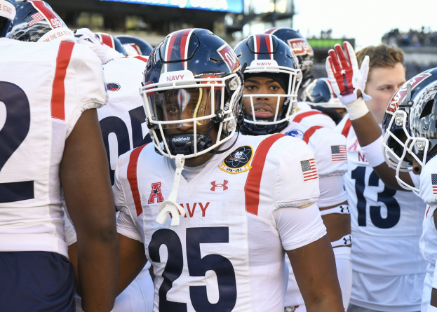 Gruppe von Männern in Fußballausrüstung auf einem Feld mit Helmen und Handschuhen, mit einer unscharfen Menge, einem Scoreboard mit der Aufschrift "Auburn vs Virginia Tech - 11/28/15 College Football Pick, Odds, and Prediction" und Himmel im Hintergrund.