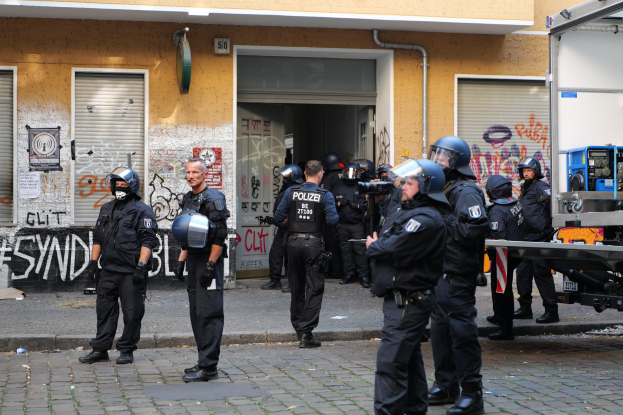 Polizisten in Helmen und Ausrüstung vor einem graffiti-bedeckten Gebäude mit einigen Waffen tragenden Beamten, einem Fahrzeug und einer Straße im Vordergrund und einem weiteren Gebäude im Hintergrund.