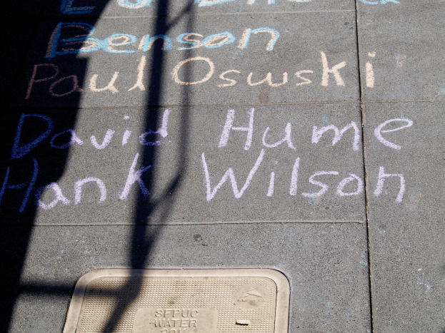 Ein Gehweg mit Kreideschrift, die wie ein Denkmal aussieht, ein Gullydeckel unten und ein Schatten eines Baums auf der linken Seite.