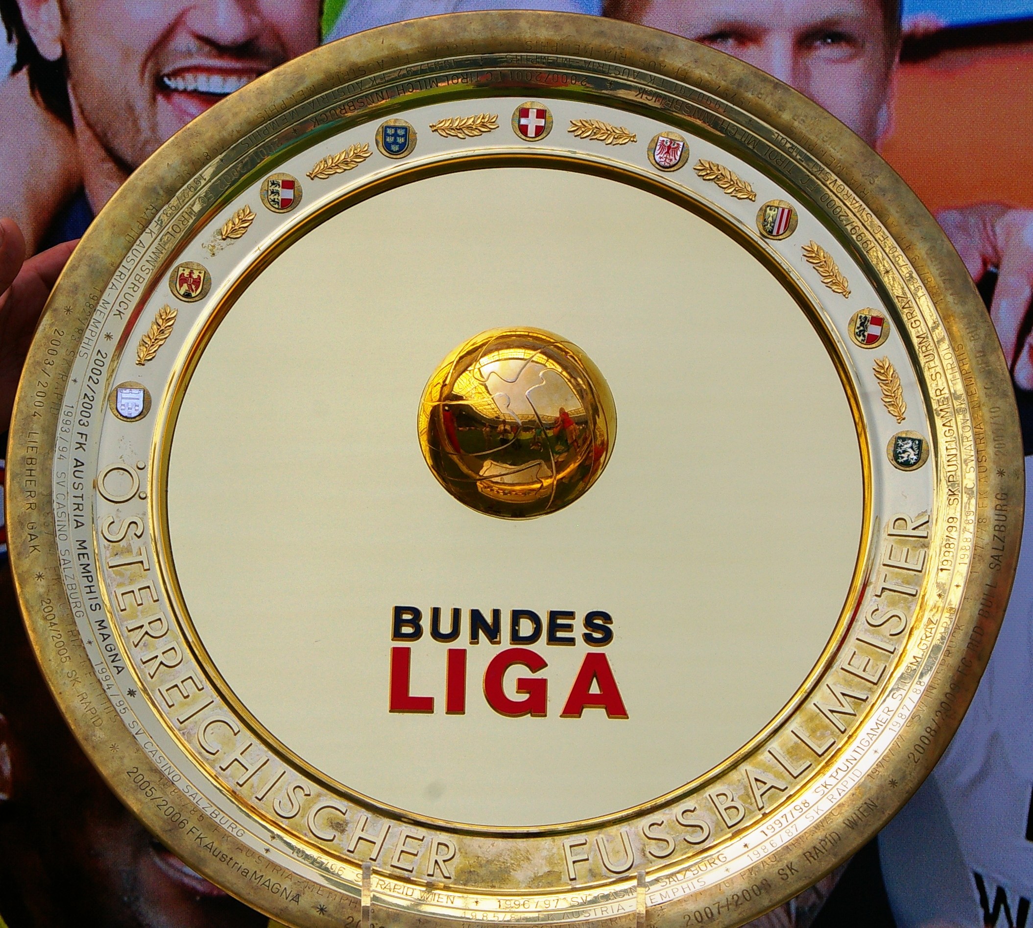 Goldener Schild mit "Bundesliga"-Text in der Hand eines Mannes, im Hintergrund ein Banner mit drei Personen.