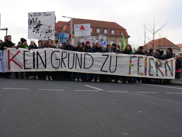 Demonstranten mit einem Banner "Kein Grund zu Feiern" gegen deutsche Sparmaßnahmen, mit Straßeninfrastruktur und Gebäuden im Hintergrund.