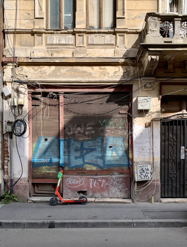 Ein roter Roller, der auf einer Straße neben einem Gebäude mit Fenstern, Rollläden und einem Tor geparkt ist, mit sichtbaren Kabeln und Straßenobjekten und Text am unteren Rand.