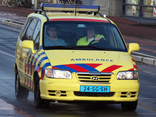 Ein gelber Krankenwagen mit zwei sichtbaren Insassen fährt an parkenden Fahrrädern auf einem Gehweg vorbei, mit einem Glasfenstergebäude im Hintergrund.