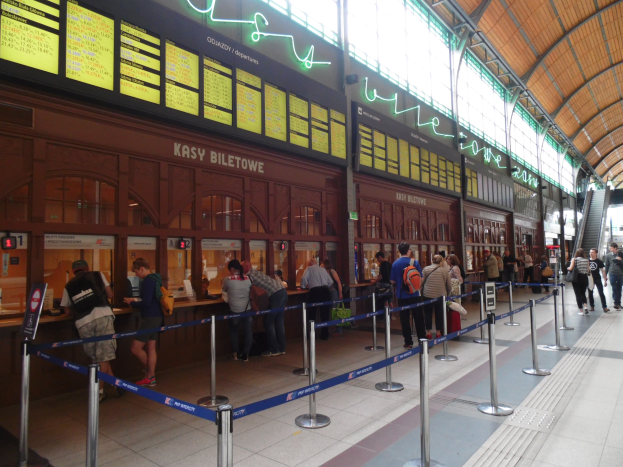 Menschen stehen in einer Reihe an einem Bahnhofsschalter mit Taschen, Stangen mit Bändern, Informationsschildern, Deckenleuchten und Treppen mit Geländern.
